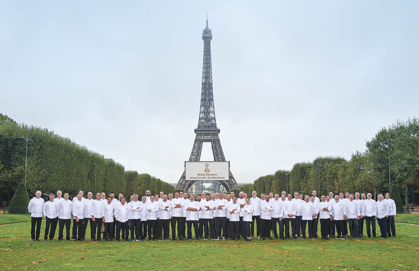 Un grand groupe de chefs vêtus d'uniformes de chef blancs, alignés en rangées sur un terrain herbeux avec la Tour Eiffel en arrière-plan et un panneau indiquant « Relais Desserts ».