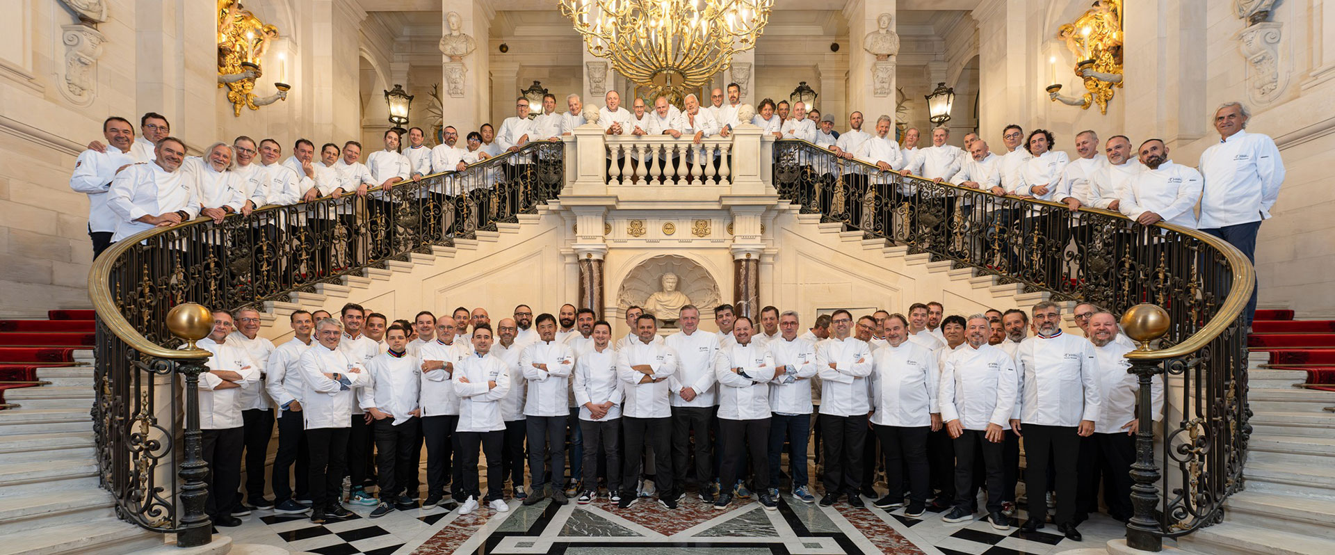 Un grand groupe de chefs en uniformes blancs debout sur un grand escalier double avec des balustrades ornées de noir et d'or, sous un lustre élaboré dans une salle luxueuse avec des sols en marbre, des statues et des candélabres muraux